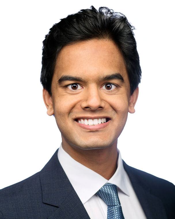 A headshot of Shomik Verma who is smiling and staring at the camera. He is wearing a suit jacket, a white collared shirt and a blue tie.