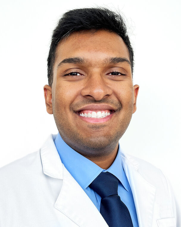 A headshot of Arjun Menta who is wearing a white doctor's coat, a blue collared shirt and a dark a blue tie.