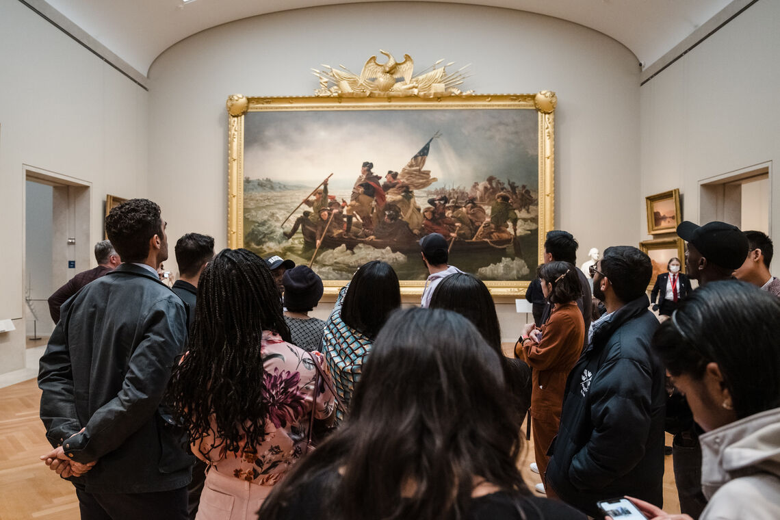 A photo of a group of Paul & Daisy Soros Fellows looking at Emanuel Leutze's painting "Washington Crossing the Delaware" which is a large-scale work in a big gold frame.