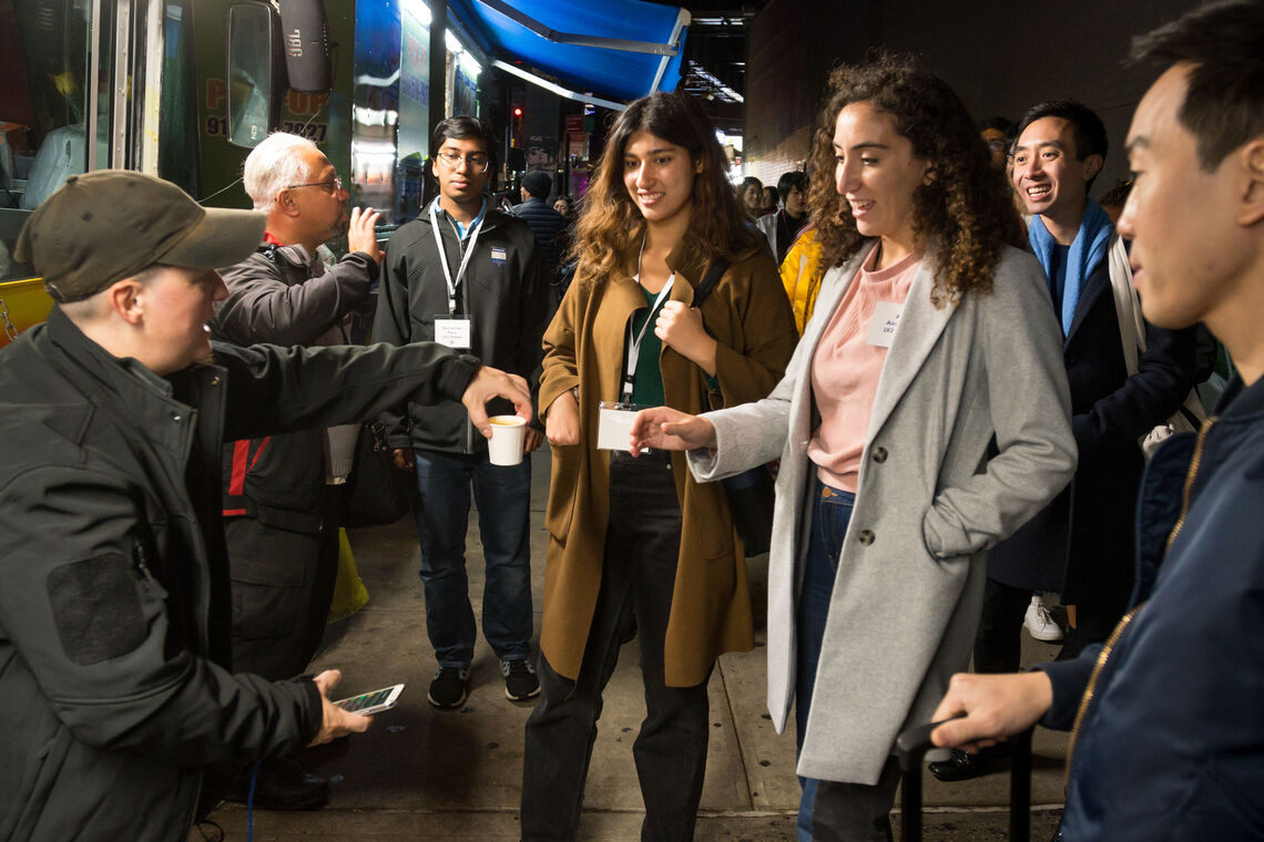 A photo taken outside at night where a group of Fellows are taking a beverage in a paper cup from a seated tour guide.